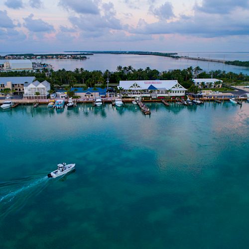 An aerial view captures a tranquil coastal town with buildings, docks, and a boat gliding across calm, clear waters under a partly cloudy sky.