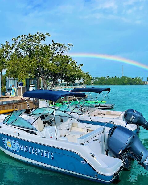 A motorboat at a dock labeled "SOLÉ WATERSPORTS" with a vivid rainbow arching over turquoise waters, surrounded by lush greenery and blue sky.