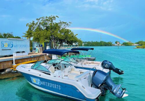 A motorboat at a dock labeled "SOLÉ WATERSPORTS" with a vivid rainbow arching over turquoise waters, surrounded by lush greenery and blue sky.