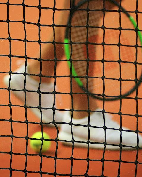 A tennis racket, ball, and player's shoes on a clay court are seen through a tennis net, which is in focus in the foreground.