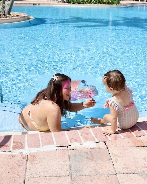 A woman and a child enjoy time by a pool, with the woman in the water and the child sitting on the edge with a toy.