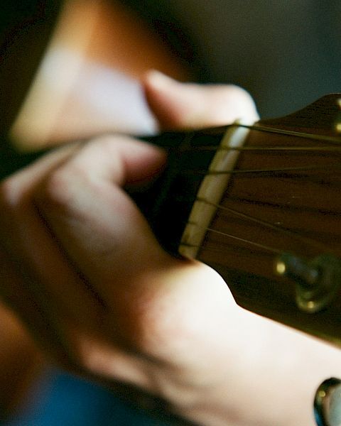 A person is playing an acoustic guitar, with a close-up view of their hand on the fretboard. The image focuses on the guitar strings and fingers.