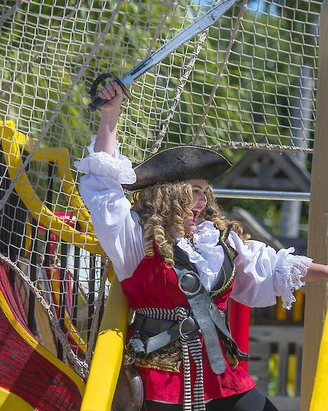 A person dressed as a pirate stands on a colorful play ship, raising a sword triumphantly while wearing traditional pirate attire.