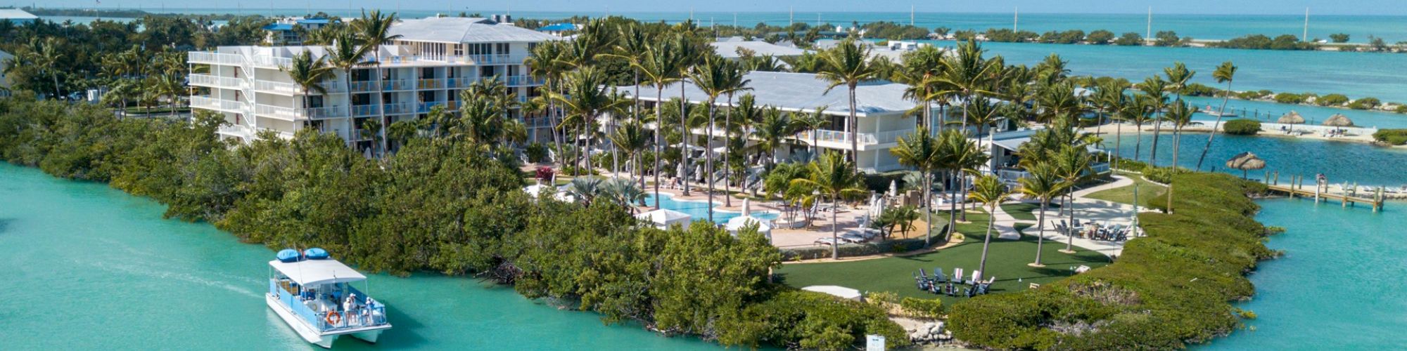 An aerial view of a resort surrounded by turquoise water, lush greenery, and a boat approaching the dock.