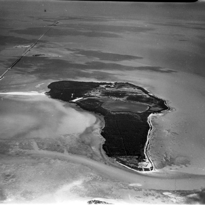 An aerial black and white photo of an isolated island surrounded by water with some land formations and a visible causeway or bridge to the mainland.