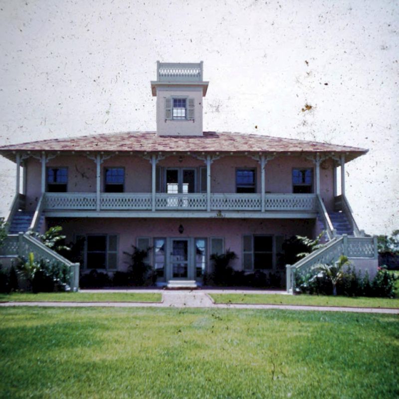 The image shows a two-story building with symmetrical stairs leading to the entrance and a cupola on the roof, surrounded by well-kept grass.