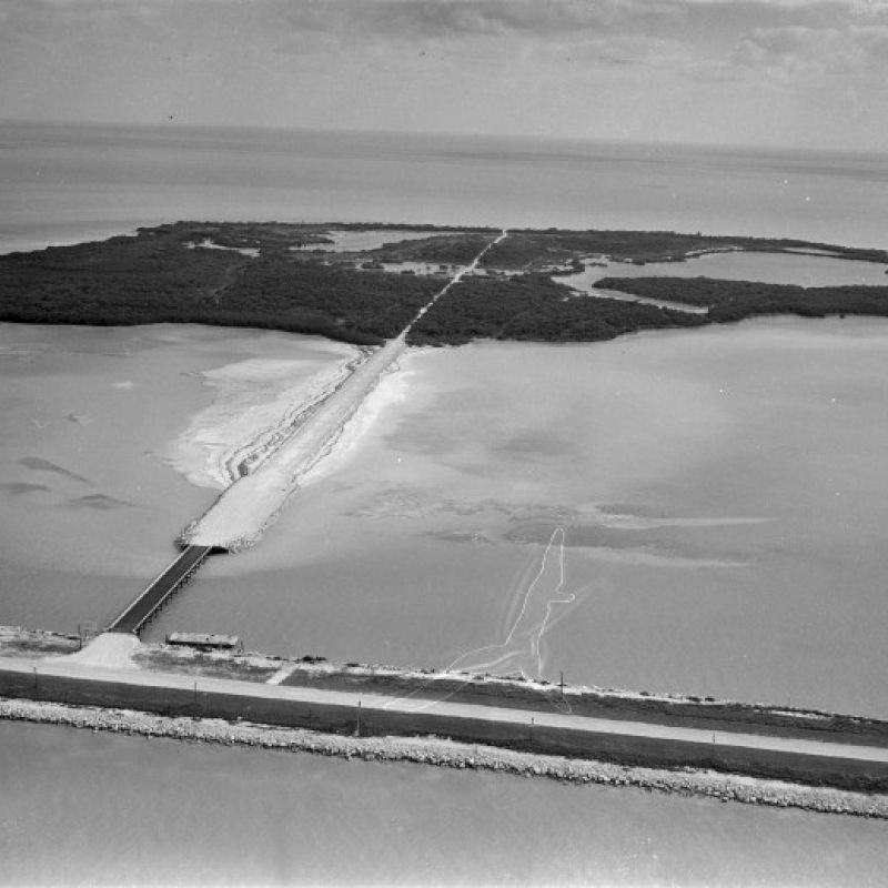 An aerial view of a causeway connecting a road to an island surrounded by water, with a bridge and sandy areas visible.