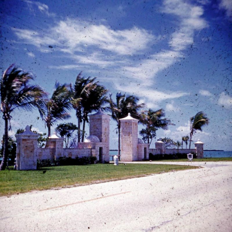 The image shows a roadside scene with tall stone gate structures, palm trees swaying in the wind, and a bright blue sky with scattered clouds.