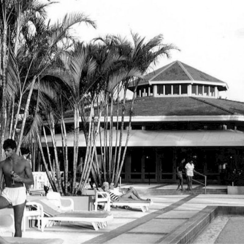 People are relaxing by a pool surrounded by palm trees in front of a round-roof building, with some sunbathing and others walking.