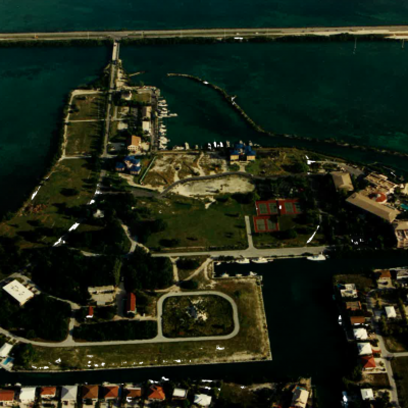 An aerial view of a coastal area with buildings, a bridge over the water, and green spaces. The area appears to be a peninsula.