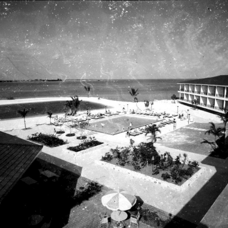 A black and white image shows a beachfront hotel with a large courtyard, central pool, palm trees, garden areas, and a view of the ocean.