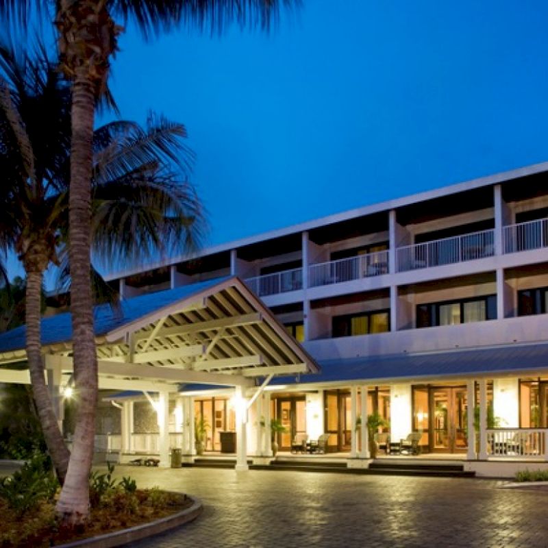 The image shows a modern hotel entrance at dusk, featuring palm trees and well-lit glass windows that create an inviting atmosphere.