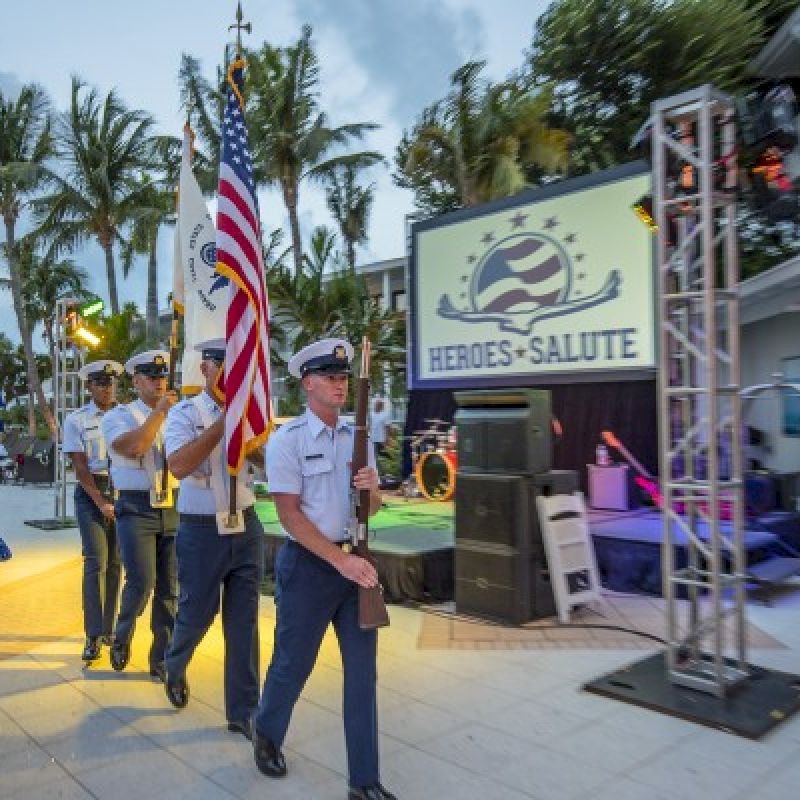 A color guard procession is taking place next to a "Heroes Salute" sign with people watching and a tropical setting in the background.