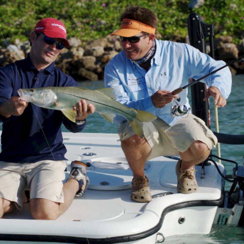 Two people on a boat are holding a large fish, smiling, and dressed in casual outdoor clothing. The boat has a Mercury motor on the back.