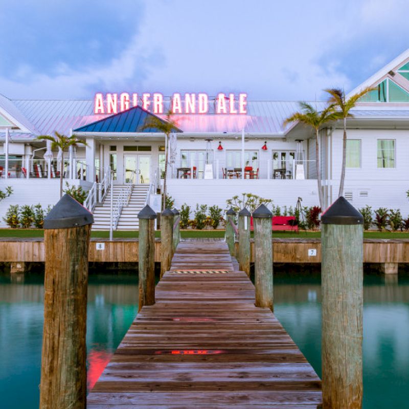 A wooden dock leads to a white building with a sign reading "ANGLER AND ALE" in pink neon, set against a backdrop of blue sky and water.