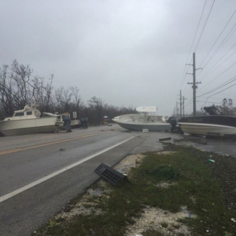 Boats are strewn across a road amidst a stormy landscape, indicative of a severe weather event or hurricane aftermath, with debris scattered around.