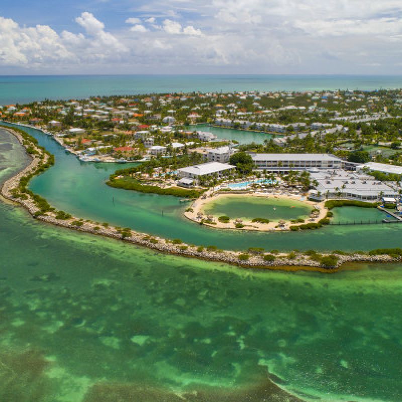 Aerial view of a tropical island with buildings, a sandy beach, and turquoise waters, surrounded by lush green vegetation and clear skies.