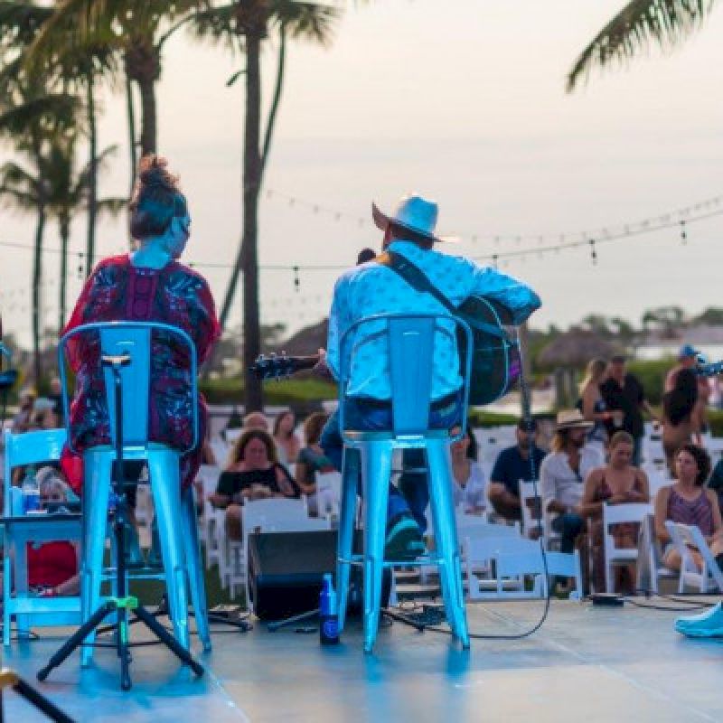 A band performs on stage outdoors with a seated audience under palm trees and string lights at sunset.