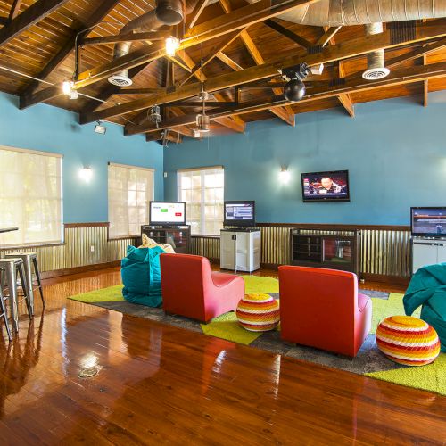 A cozy lounge with vibrant chairs, bean bags, bar stools, and a few mounted TVs, featuring wooden floors and a high, exposed-beam ceiling.