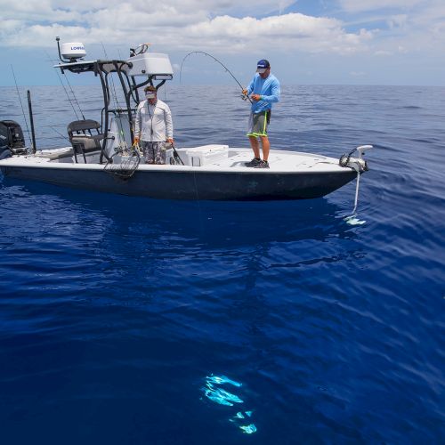 Two people on a small boat fishing in a vast ocean with clear, blue waters; one person is holding a fishing rod that appears to have caught a fish.