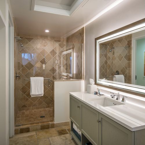 This image shows a modern bathroom with a large mirror, double sink vanity, and a walk-in tiled shower with a towel hanging on the glass door.