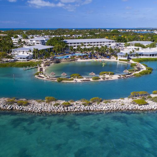 An aerial view of a tropical resort with buildings, palm trees, a beach area, and surrounding water, featuring a boat in the foreground.