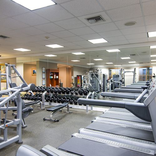 The image shows an empty gym with various equipment, including treadmills, weight machines, and dumbbells, under a well-lit ceiling.