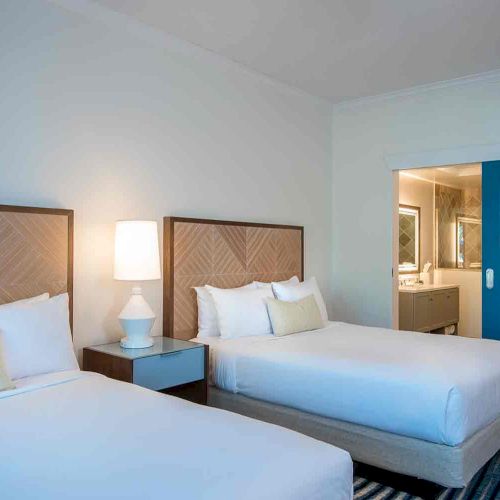 A room with two single beds, a bedside table with a lamp, a blue door leading to a bathroom with a vanity mirror.