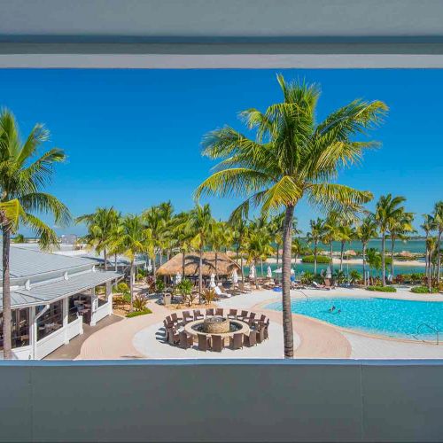 The image shows a scenic resort with a pool, palm trees, outdoor seating around a fire pit, and a view of clear blue skies and water.