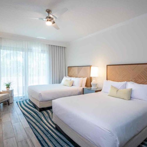 A modern hotel room features two queen beds, a seating area, a ceiling fan, and well-lit decor with light wood and white elements.