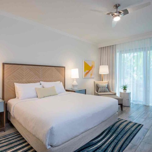 A modern bedroom with a large bed, two side tables with lamps, an armchair, a ceiling fan, a striped rug, and a large window with sheer curtains.