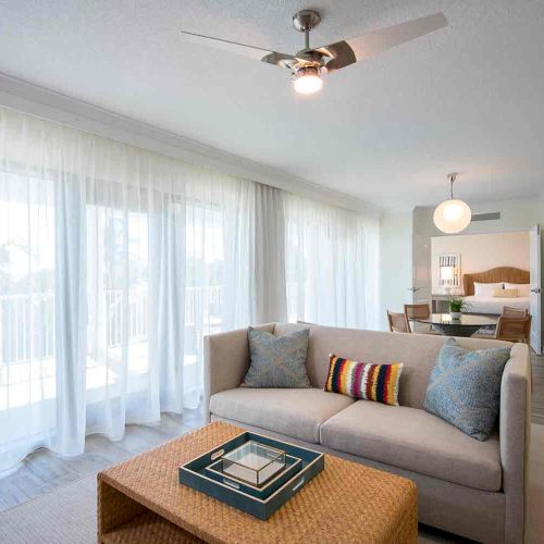 A bright, modern living room with floor-to-ceiling windows, sheer curtains, a beige sofa, colorful pillows, a coffee table, and an adjoining dining area.