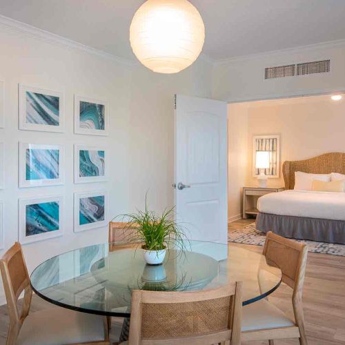 A modern, neatly decorated room with a glass dining table, four chairs, wall art, and an adjacent bedroom featuring a bed with a rattan headboard.