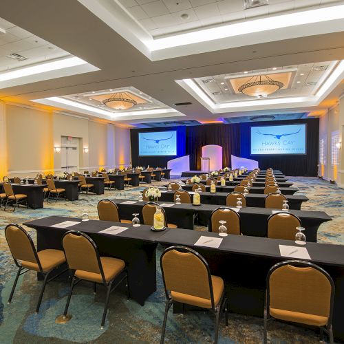 A conference room is set up with rows of tables and chairs facing a stage with two large screens, prepared for a presentation or event.