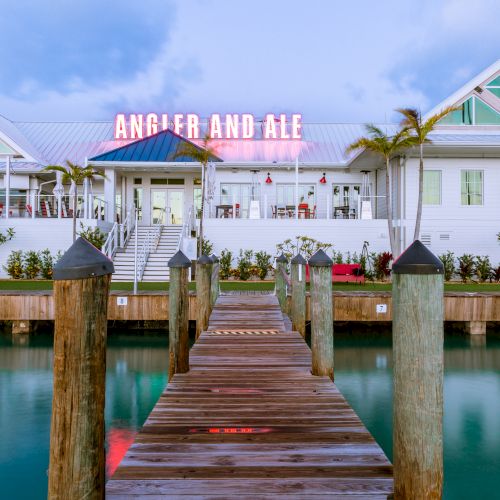 A wooden dock leads to a white building with "Angler and Ale" in neon lights, surrounded by palm trees and reflected in clear blue water.