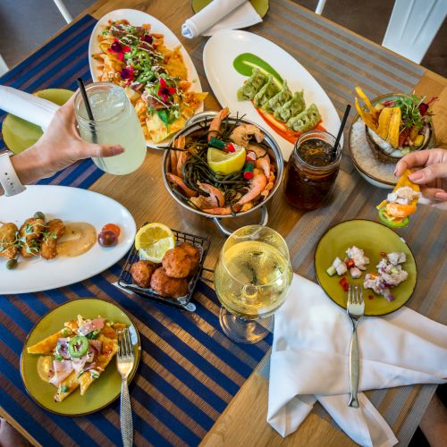 A table with various seafood dishes, appetizers, and drinks being enjoyed by two people. One holds a drink, while the other holds a shrimp.