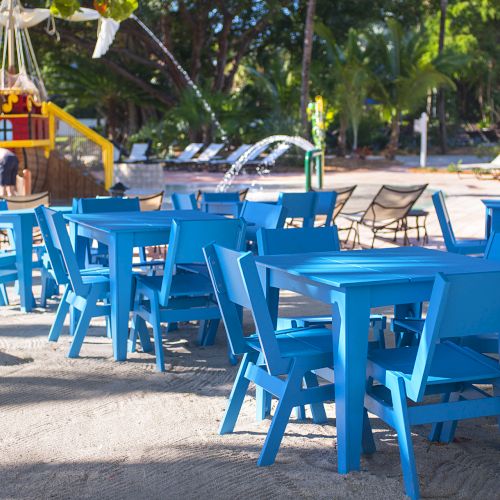 A group of bright blue tables and chairs is set up on sandy ground near a playground and trees in the background.