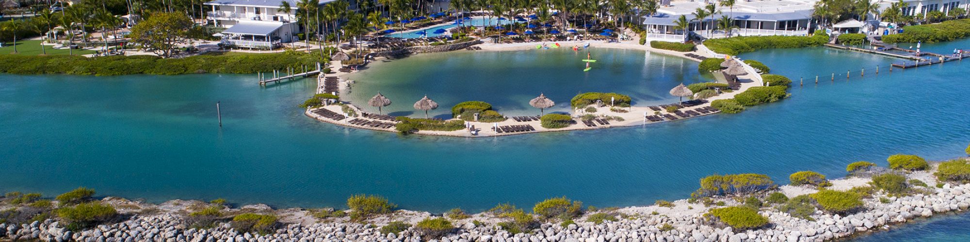 An aerial view of a coastal resort with white buildings surrounded by water and greenery, featuring a small sandy beach area and a boat on the water.