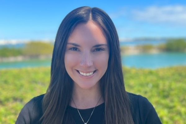 The image shows a woman with long, dark hair smiling at the camera, with a scenic background of water and greenery under a clear blue sky.