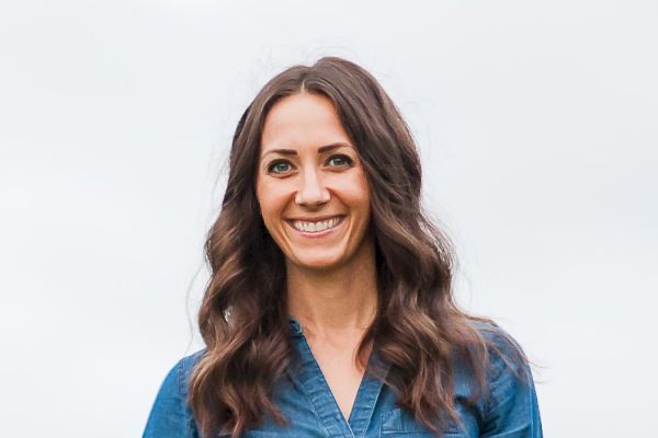 The image shows a smiling woman with long, wavy hair, wearing a denim dress held by a brown belt, standing against a light, cloudy sky background.