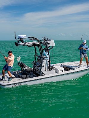 Two people are fishing on a boat in a large body of water with clear skies. Both are holding fishing rods and appear to be reeling in catches.