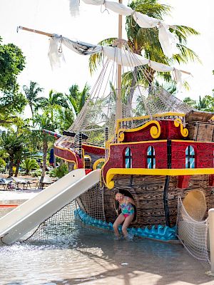 A child plays on a pirate-themed water slide in a pool area with palm trees in the background, featuring a colorful ship structure.