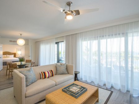 A modern living room with a sofa, coffee table, large windows with sheer curtains, and a connected bedroom in the background.