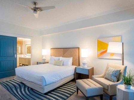 A modern bedroom with a bed, two nightstands, a patterned area rug, an armchair, a lamp, and abstract art on the wall, leading to a bathroom.