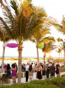 A group of people is gathered at a beachside event under palm trees, with colorful lanterns decorating the area and a scenic ocean view.