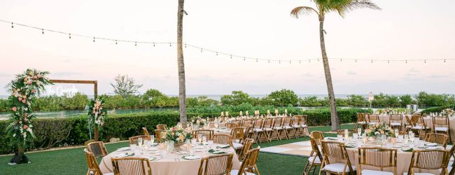 An outdoor event setup with round tables, chairs, string lights overhead, and palm trees in the background, likely for a wedding or celebration.