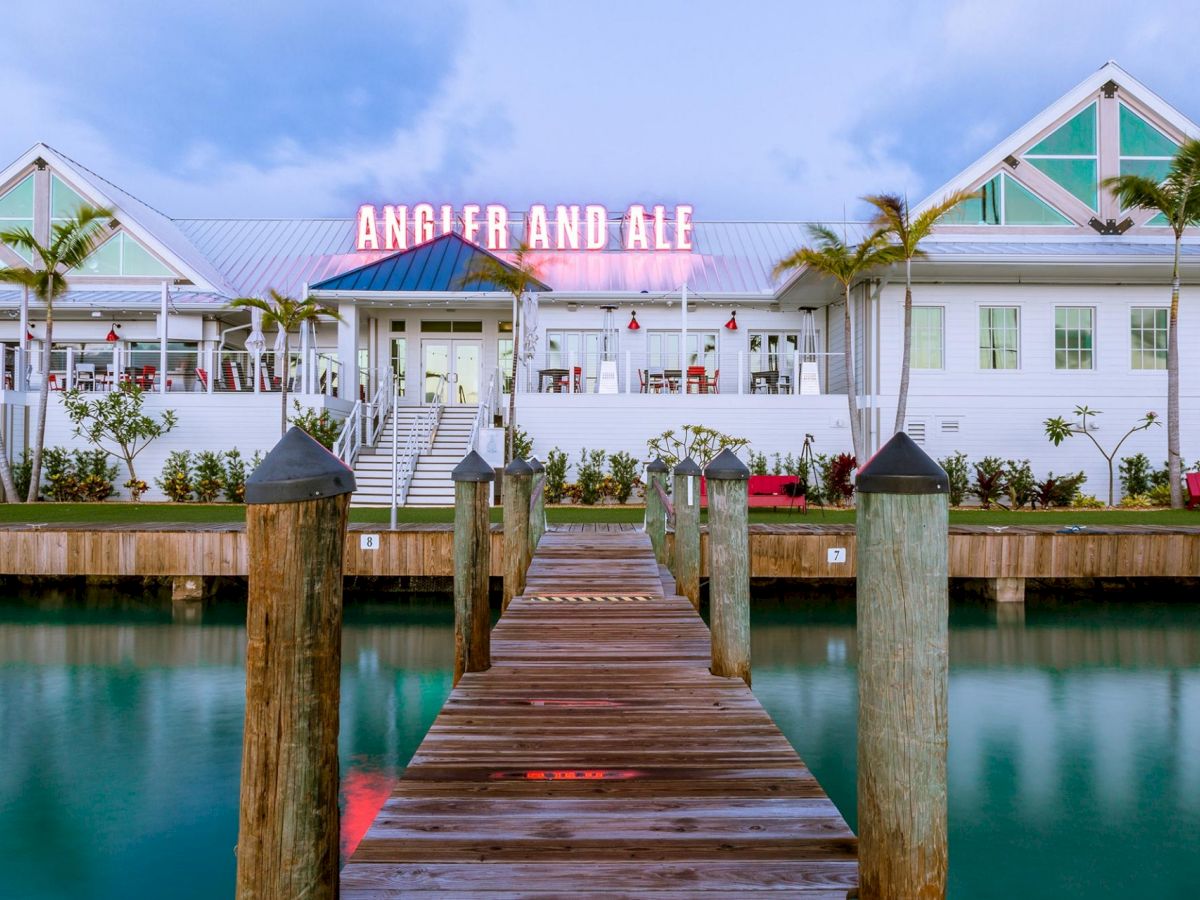 A wooden dock leads to a waterfront building with a neon sign that reads "ANGLER AND ALE," surrounded by palm trees and calm water.