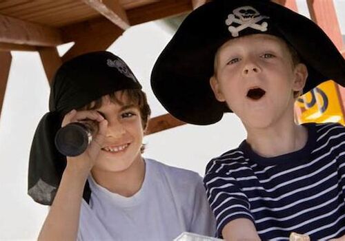 Two children are dressed as pirates, one using a telescope and the other with an open mouth, both wearing pirate hats.