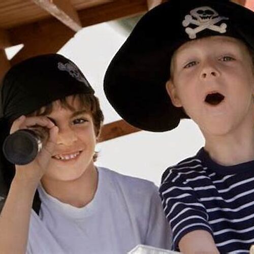 Two children are dressed as pirates, one using a telescope and the other with an open mouth, both wearing pirate hats.