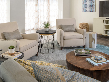 A cozy living room with beige armchairs, a round wooden coffee table, a TV on a console, and books and decor, all on a patterned rug.